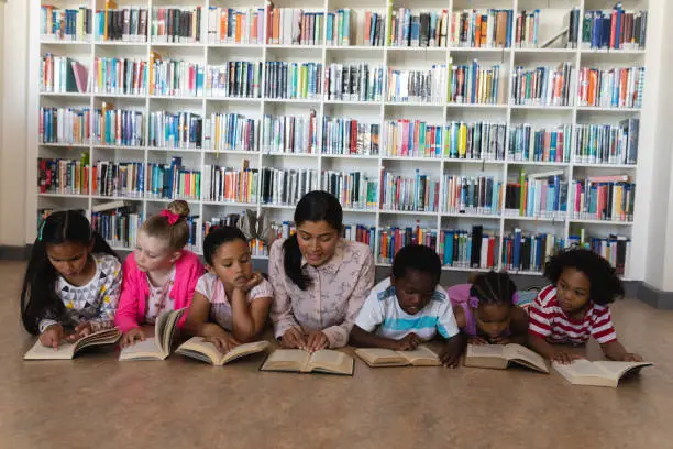 Front view of female teacher and schoolkids reading a book while lying on floor of school library