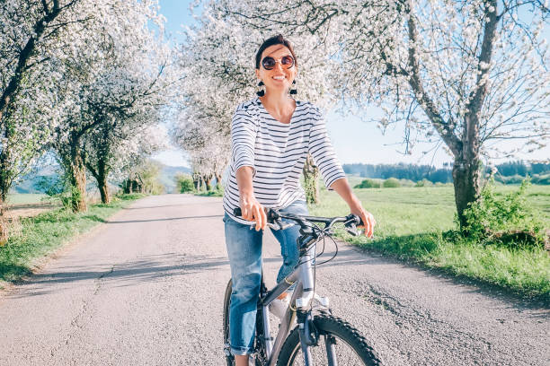 feliz sonriente mujer monta una bicicleta en la carretera bajo los árboles de flor. primavera viene imagen de concepto. - comming fotografías e imágenes de stock