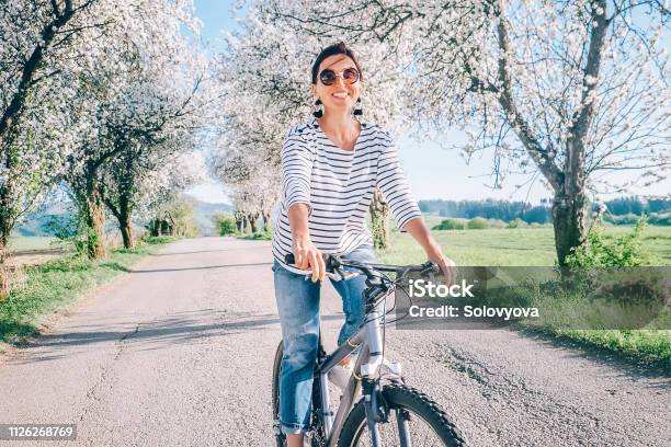 幸せな笑顔の女性の花の木の下で田舎道を自転車に乗る春にはコンセプト イメージが来ています - 春のストックフォトや画像を多数ご用意 - 春, 女性, サイクリング