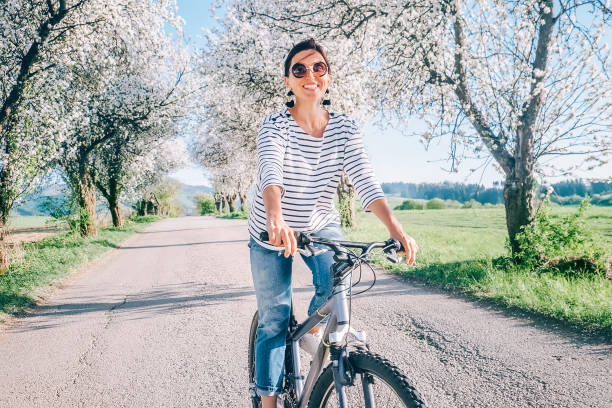 szczęśliwa uśmiechnięta kobieta jeździ na rowerze po wiejskiej drodze pod kwitnącymi drzewami. nadchodzi wiosna. - nature smiling teenage girls female zdjęcia i obrazy z banku zdjęć