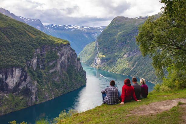 People looking at the Geirangerfjord, Norway Four people are sitting on the ground and looking at the Geirangerfjord on a cloudy summer day. The rocky mountains are covered with trees. geirangerfjord stock pictures, royalty-free photos & images