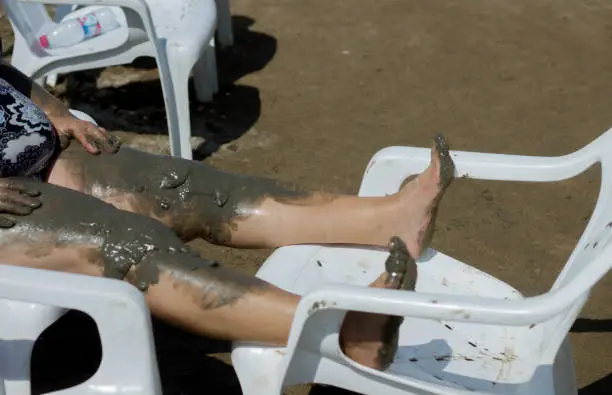 Middle aged woman donning retro looking swim suit with Dead Sea mud on her stretched out legs resting on chair under the sun