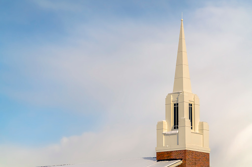 Snowy church steeple and roof against cloudy sky. Close up a church's steeple and snow covered roof in Eagle Mountain, Utah. Scenic winter landscape with a bright cloudy blue sky in the background.