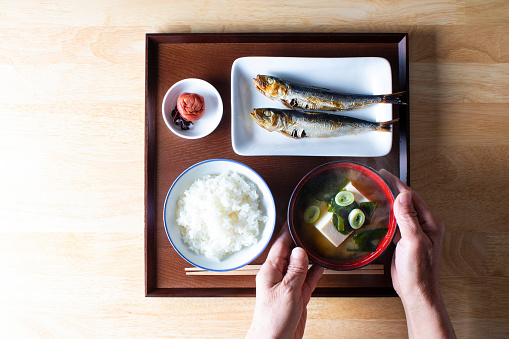 The menu is cooked rice, baked sardines, tofu and seaweed miso soup and Umeboshi(plum). senior Japanese woman with a bowl of miso soup.