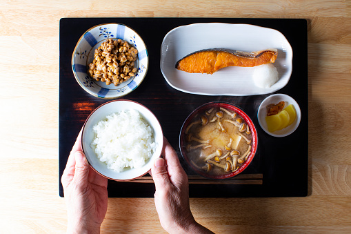 The menu is cooked rice, baked salmon, Natto, Nameko (mushrooms) and tofu miso soup and Tkuan (pickle of radish). senior Japanese woman's hand with a bowl full of cooked rice.