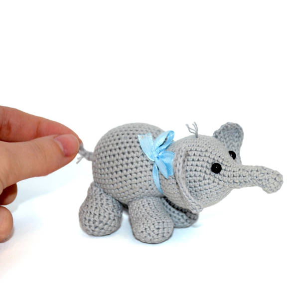 istock Isolated crocheted gray elephant with a blue bow on the neck on a white background. Female hand holds the elephant by the tail 1126362933