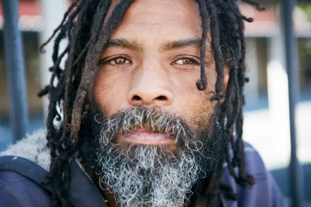A weather beaten homeless person looks unhappy, standing outdoors with dreadlocks and a graying beard.