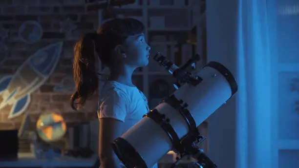 Cute young girl stargazing at night with a telescope, she is looking away: imagination and childhood concept