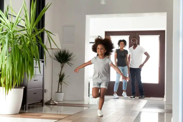 Photo of Excited little funny african girl running exploring big modern house on moving day