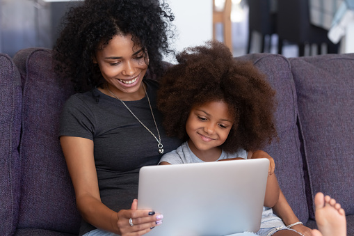 Smiling african american mother and kid daughter having fun with computer sitting on couch, happy mixed race mom with child girl using laptop, watching cartoons, making video call, doing shopping