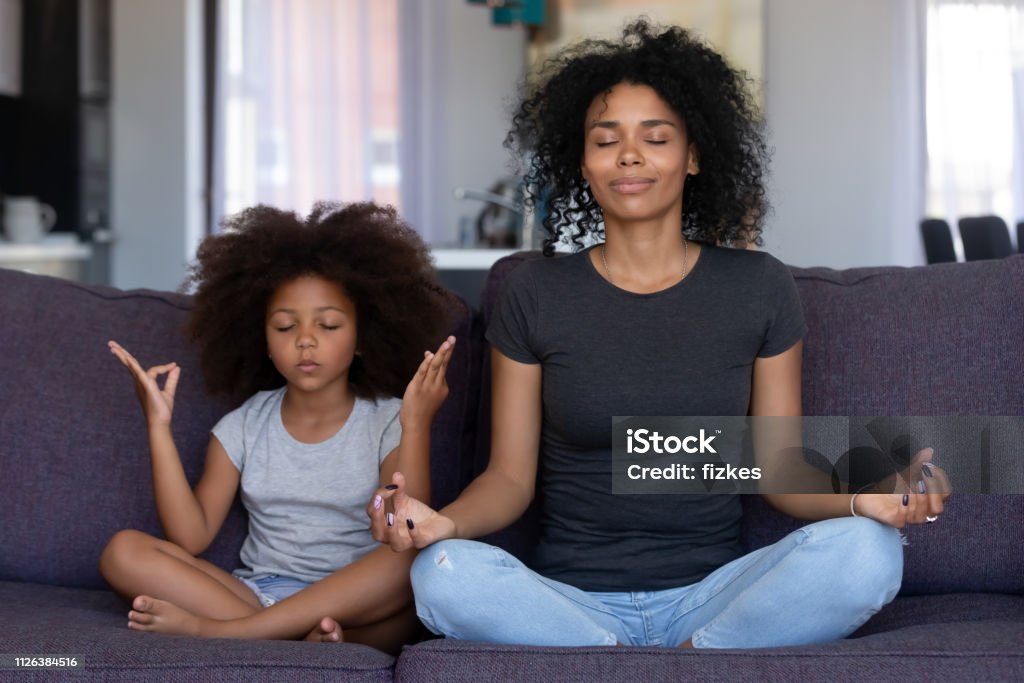 Mindful african mom with funny kid daughter doing yoga together Mindful african mom with cute funny kid daughter doing yoga exercise at home, calm black mother and mixed race little girl sitting in lotus pose on couch together, mum teaching child to meditate Child Stock Photo