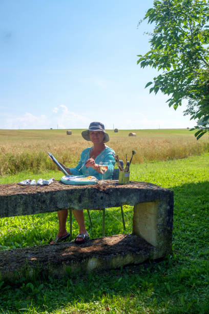 An artist sits with a rural countryside view painting A lady artist sits in late afternoon shade with a rural countryside view working on the painting she started earlier in the day. painted image paintings oil paint senior women stock pictures, royalty-free photos & images