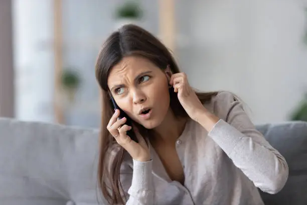 Annoyed young woman talking on the phone not hearing bad signal lost no mobile connection on cellphone, irritated girl closing ear frustrated by communication telecommunication service problem indoor