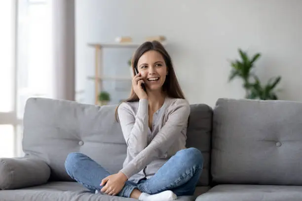 Happy young woman caller talking on the phone at home, cheerful teen girl enjoys pleasant mobile conversation, smiling millennial female holding cell speaking making call by telephone in living room
