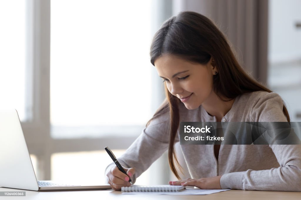 Young woman writing notes in notebook making list planning tasks Young professional woman intern student noting in notebook studying at home office, teen girl writing essay or preparing for test exam with laptop, copywriter freelancer making list planning tasks Writing - Activity Stock Photo