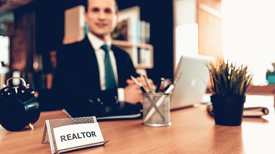 Real Estate Agent is Looking to Camera and Smiling. Real Estate Agent is Caucasian Adult Man. Man is Sits at Desk. Person is Wears Business Suit. Person in Office. Sunny Daytime. Real Estate Agent Sign on Table. Blur View of Man.