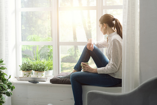 Young woman relaxing at home next to a window and having a cup of coffee