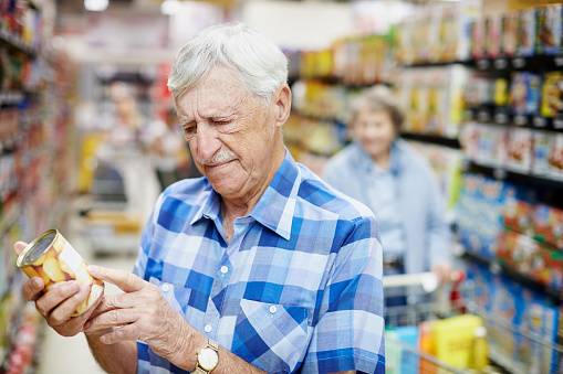 A senior man in a supermarket looks down at the label of a product he holds, checking it seriously as his wife follows behind him.