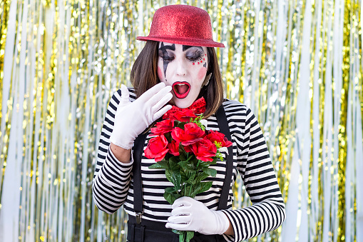 Woman dressed as a mime. dressed in a black and white striped shirt, black suspenders, a red shirt and a hat, with black pants. Looking up holding the hat. Holding a bouquet of roses with surprise face.