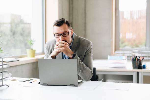 Thoughtful middle aged handsome businessman in shirt working on laptop computer in office stock photo