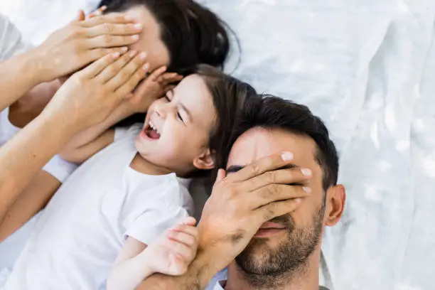 Photo of Top view of beautiful family, pretty mother, handsome father and cute kid playing outdoors in park. Horizontal portrait of female and male closed eyes with hand play peek a boo with child. Parenting