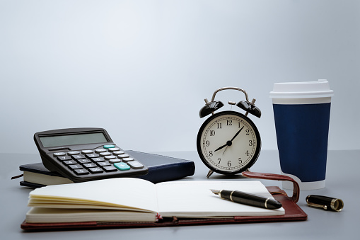 alarm clock with notebook, calculator, pen and cup of coffee on gray background on working table