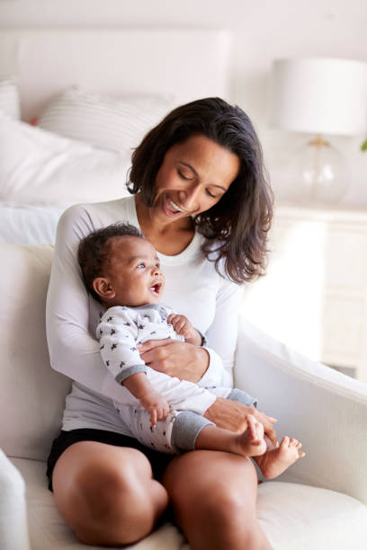 Young adult mother sitting in an armchair in her bedroom, holding her three month old baby son in her arms and looking down at him smiling, vertical Young adult mother sitting in an armchair in her bedroom, holding her three month old baby son in her arms and looking down at him smiling, vertical biracial newborn stock pictures, royalty-free photos & images