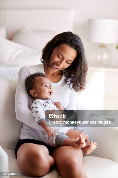 Young Adult Mother Sitting In An Armchair In Her Bedroom Holding Her Three Month Old Baby Son In Her Arms And Looking Down At Him Smiling Vertical Stock Photo - Download Image Now
