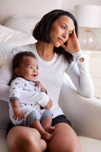 Young adult mother sitting in an armchair in her bedroom, holding her three month old baby son, looking away in contemplation, vertical Young adult mother sitting in an armchair in her bedroom, holding her three month old baby son, looking away in contemplation, vertical postpartum depression stock pictures, royalty-free photos & images