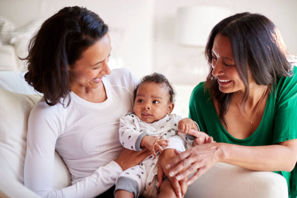 madre millenaria di razza mista seduta su una poltrona che tiene il figlio di tre mesi, sua madre inginocchiata accanto a loro - domestic life mother baby multi ethnic group foto e immagini stock