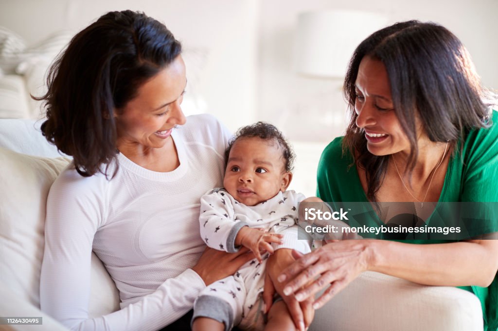 Millennial mixed race mother sitting in an armchair holding her three month old baby son, her mother kneeling beside them Baby - Human Age Stock Photo