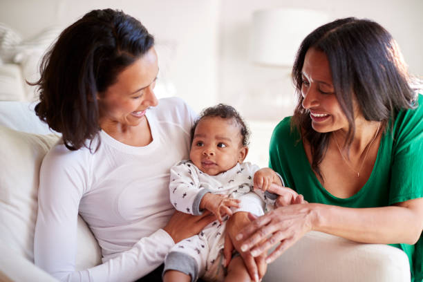 Millennial mixed race mother sitting in an armchair holding her three month old baby son, her mother kneeling beside them Millennial mixed race mother sitting in an armchair holding her three month old baby son, her mother kneeling beside them lounge chair photos stock pictures, royalty-free photos & images