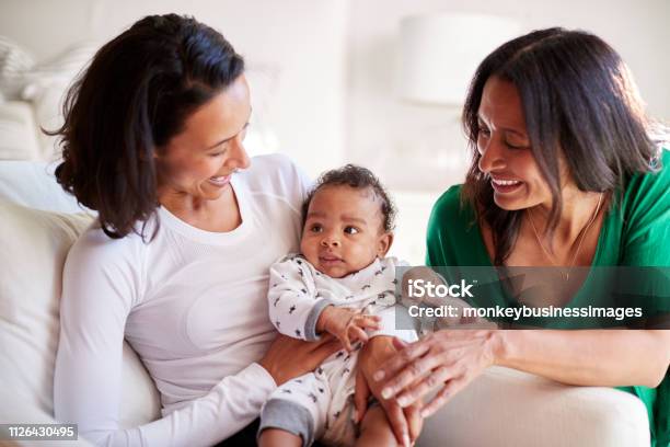 Millennial Mixed Race Mother Sitting In An Armchair Holding Her Three Month Old Baby Son Her Mother Kneeling Beside Them Stock Photo - Download Image Now