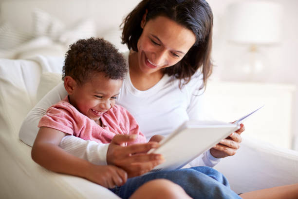 gros plan de race mixte jeune adulte mère assis dans un fauteuil, lisant un livre avec son fils âgé de trois ans sur ses genoux, riant, gros plan - family reading african descent book photos et images de collection