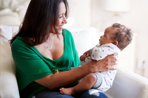 avó de raça mista envelhecido médio sentado em uma poltrona segurando seu neto de três meses de idade e sorrindo, concentre-se em primeiro plano - baby grandparent newborn grandmother - fotografias e filmes do acervo