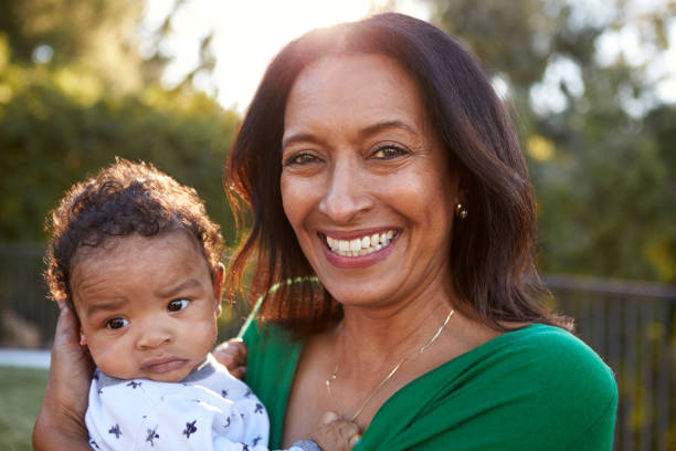 happy mixed race middle aged grandmother holding her three month old grandson in the garden and smiling to camera, close up, head and shoulders - babies and children close up horizontal looking at camera imagens e fotografias de stock