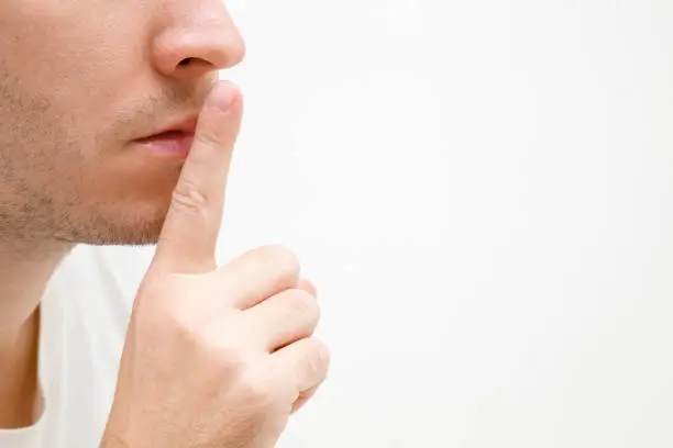 Young man's finger showing shush gesture. Sign for silence. Face closeup. Isolated on white background. Empty place for text, quote or sayings.