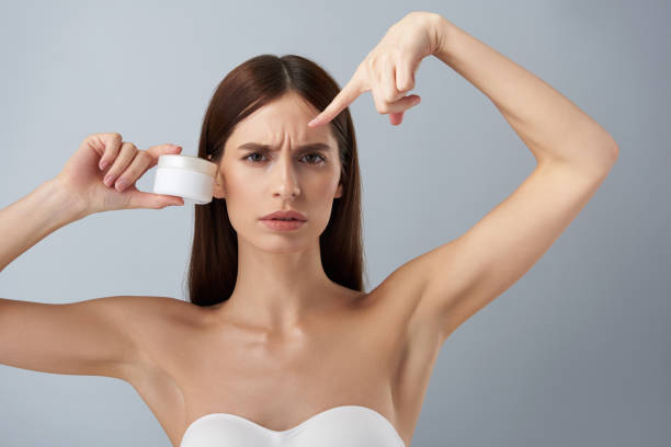 Annoyed young woman with cosmetic product showing skin problems on her forehead Close up portrait of beautiful brunette girl in white bra holding jar of cream while pointing finger at wrinkles on her forehead. She frowning and looking at camera with irritated expression wrinkled forehead stock pictures, royalty-free photos & images