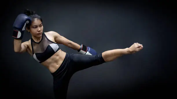Photo of Young Asian woman boxer with blue boxing gloves kicking in the exercise gym, Martial arts on black background. Female boxing class concept
