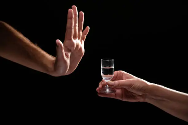 Studio shot black backgroud man's hand in the gesture of refusal vs woman's palm of hand passing glass of alcohol
