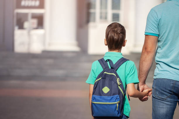 Father leads a little child school boy go hand in hand. Parent and son with backpack behind the back. stock photo