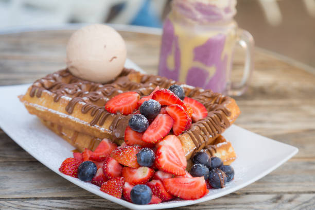Close up Blegium waffles topped with strawberry, blueberry, ice cream ball and chocolate sauce served with milkshake on wooden table on a hot summer day date stock photo