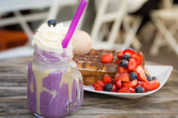 Close up blueberry milkshake with whipped cream and berry on top with Blegium waffles topped with strawberry, blueberry, ice cream ball and chocolate sauce served on wooden table on a hot summer day date stock photo