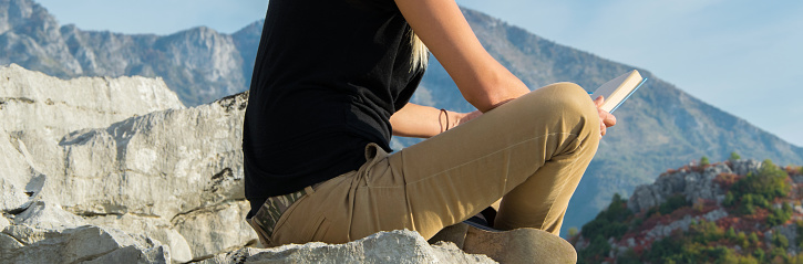 Young blond woman sitting on the edge of the mountain  cliff reading a book against beautiful mountains peak. Healthy lifestyle/travel concept