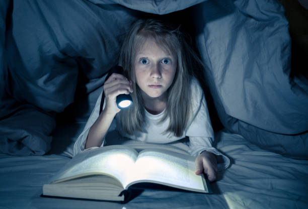 Beautiful caucasian girl lying in bed under the duvet holding a lantern reading a mystery book in the dark late at night looking frightened with a dramatic mood light. stock photo