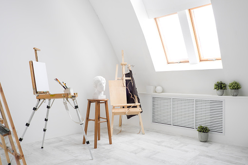 White and bright studio with a window and gray floor. Workspace of the artist. Easel, canvases and plaster figures for learning to draw. Conceptual interior without people.