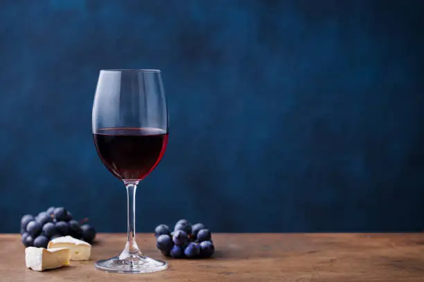 Photo of Glass of red wine with fresh grape and cheese on wooden table. Blue background. Copy space.