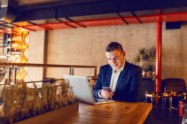 istock Middle aged businessman dressed smart casual using smart phone and sitting in cafeteria. In front of him on a desk laptop. 1126494273