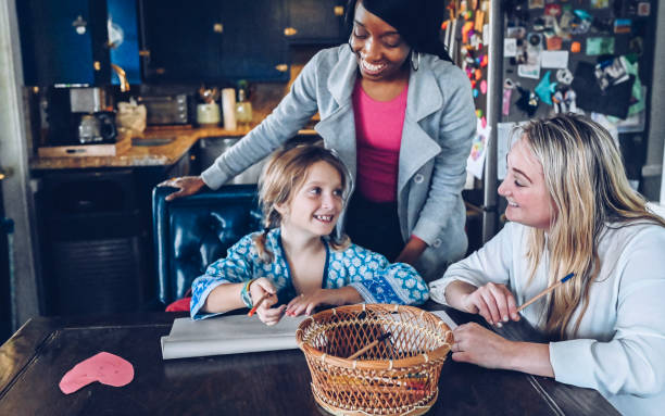 Same sex couple help child with homework at home Mixed ethnicity female same sex lesbian couple help child with homework. Little girl is adorable elementary age student and has paperwork and art supplies in front of her. Working at the kitchen table in a domestic home. Typical weekday routine for a modern family, cheerful, helpful and togetherness gay dinner table stock pictures, royalty-free photos & images