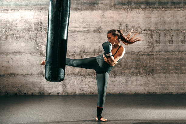 bruna forte dedicata con coda di cavallo, abbigliamento sportivo, piede nudo e con guanti da boxe che calciano sacco in palestra. - arte marziale foto e immagini stock